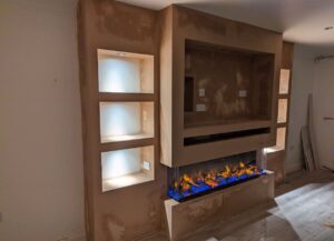 Corner media wall with recessed TV space, illuminated shelving niches, built-in soundbar slot, and a wide electric fireplace featuring blue and orange LED flames.