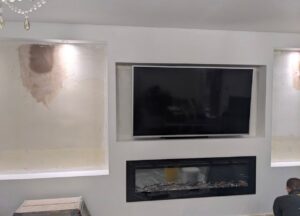 White media wall with recessed TV, illuminated display alcoves on both sides, and a built-in electric fireplace in a living room undergoing finishing work.