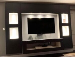 Modern media wall with a mounted TV, built-in illuminated display niches, overhead spotlights, and a floating shelf unit in a dark contemporary living room design.