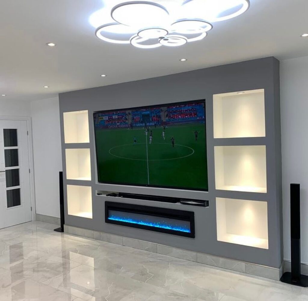 Modern grey media wall with large mounted TV, illuminated display alcoves on both sides, built-in soundbar shelf, and electric fireplace featuring blue LED flames in a contemporary living room.