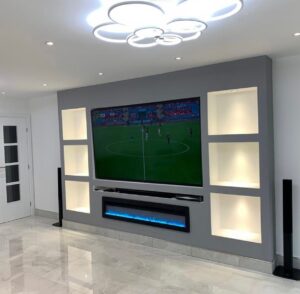 Modern grey media wall with large mounted TV, illuminated display alcoves on both sides, built-in soundbar shelf, and electric fireplace featuring blue LED flames in a contemporary living room.