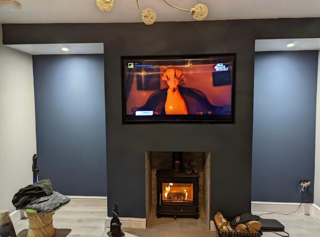 Dark feature wall with mounted flat-screen TV and a built-in wood-burning stove surrounded by stone detailing, set between two recessed alcoves with spotlights in a modern living room.