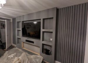 Grey modern media wall with recessed TV space, multiple built-in shelving niches, vertical slat wall panels, and an unfinished section for an electric fireplace in a contemporary living room.